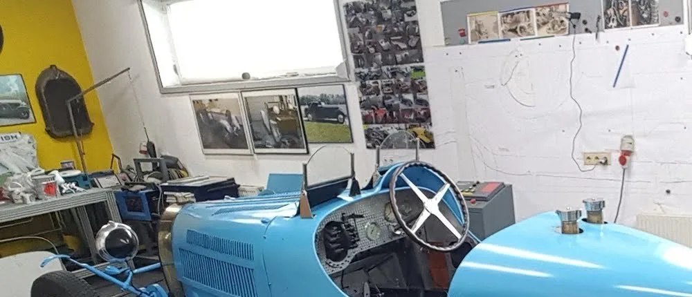 Blick in die Werkstatt: ein hellblauer Vorkriegs-Rennwagen steht auf der Hebebühne, an der Wand hängt eine Karosseriezeichnung in Originalgröße.
